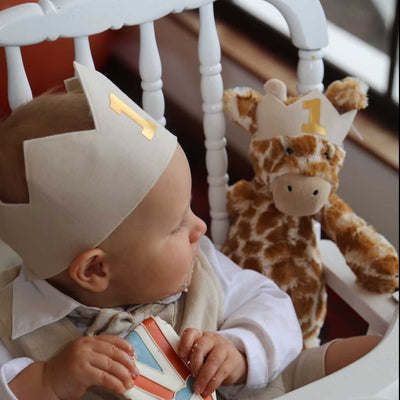 baby boy sits in high chair eating a cookie for his first birthday photoshoot. He is wearing a first birthday crown with a gold 1 on the front. He sits next to his toy giraffe who also wears a matching toy birthday crown