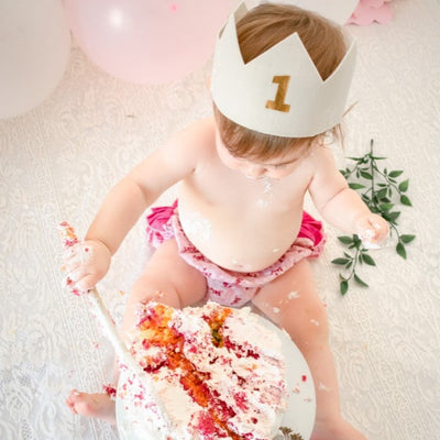 cake smash photoshoot for a girls first  birthday. 1 year old girl is holding a spoon smashing cake with pink romper and a white first birthday crown