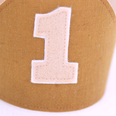 close up of a tan number 1 made from felt, stitched onto the front of a linen first birthday crown in caramel brown