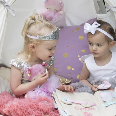 2 young girls read a book sittingin a teepee surrounded by pillows and soft toys they are wearing a tutus. One girls wears a silver glitter tiara and the other a white bow in her hair