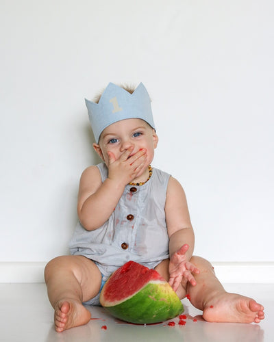 1 year old boy wearing a blue linen birthday crown and a pale blue romper eating watermelon for a one in a melon themed first birthday photoshoot