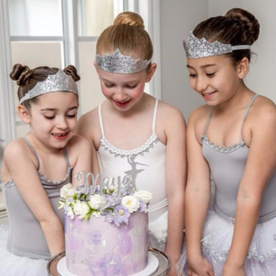3 young girls around a birthday cake topped with white and purple flowers, dressed in ballet tutu and wearing a silver glitter tiara crown each