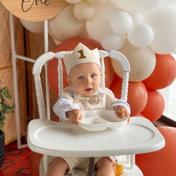 First birthday boy sits in a highchair with balloons in the background. He is dressed in a white shirt, bow tie and white linen birthday crown with a gold 1 on the front. He is holding out a tray for some food