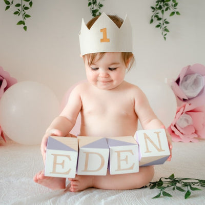 1  year old girl sits for her cake smash photoshoot  session with wooden blocks in her hands. There are paper flowers and vines in the background. She wears a handmade white linen first birthday crown with a gold 1 on the front. 